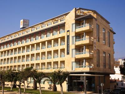 Modernes sechsstöckiges Hotelgebäude mit Balkonen und Bäumen davor bei klarem Himmel.