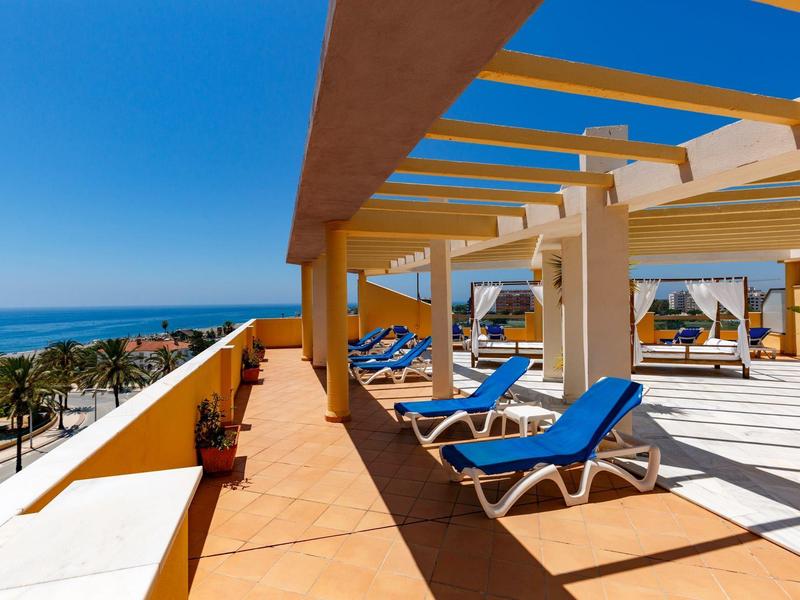 Terrassenbereich mit blauen Liegestühlen, Holzpergola und Blick auf Meer und Strand bei klarem Himmel.