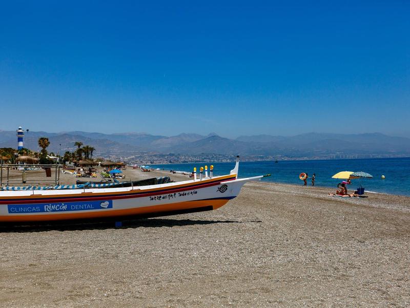 Ein bunter Fischerboot liegt am Sandstrand, blauer Himmel und ruhiges Meer mit Menschen im Hintergrund.