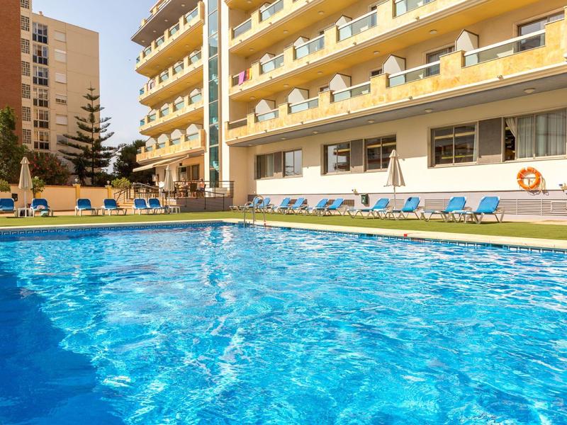 Schwimmbad mit klarem Wasser vor gelbem Gebäudekomplex und blauen Liegestühlen.