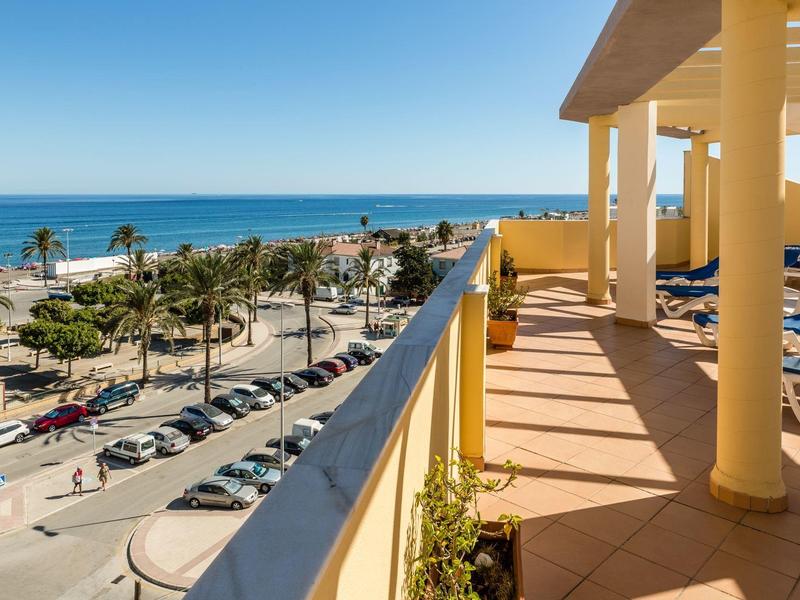 Balkon mit Säulen, Liegestühlen und Meerblick, Palmen und Autos an der Straßenpromenade.