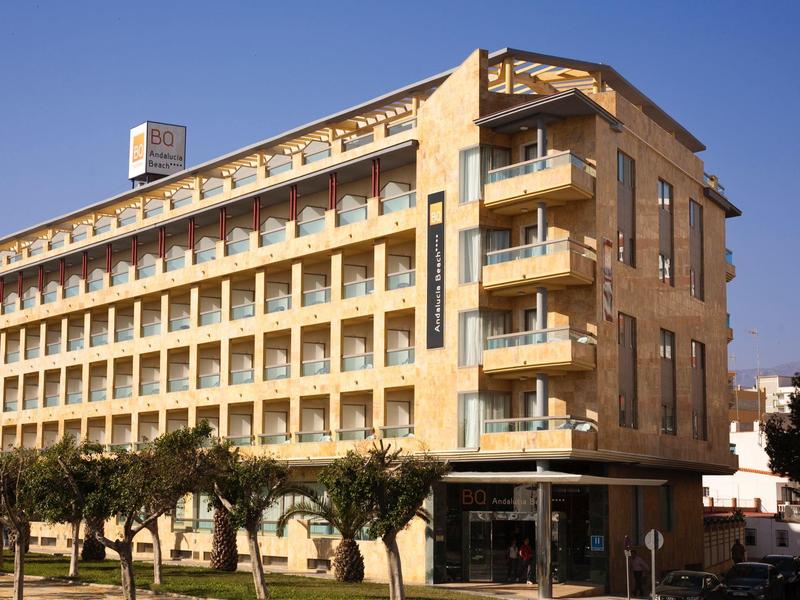 Modernes sechsstöckiges Hotelgebäude mit Balkonen und Bäumen davor bei klarem Himmel.