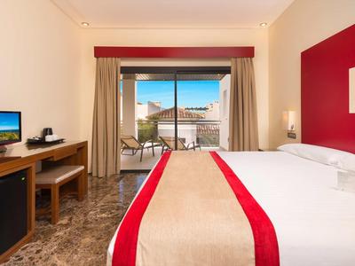 Hell eingerichtetes Hotelzimmer mit großem Bett, Fernseher und Balkon mit Sitzgelegenheit.