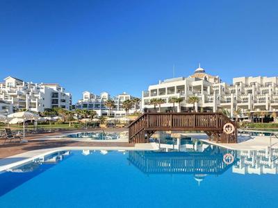 Großer blauer Pool vor weißen Hotels unter klarem Himmel mit Holzbrücke und Palmen.