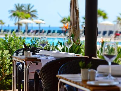 Restaurantterrasse mit gedeckten Tischen, Blick auf Palmen, Strandliegen und das blaue Meer im Hintergrund.