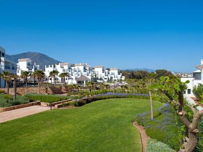 Grüner Garten mit Wegen, weißen Häusern und Bergen im Hintergrund unter blauem Himmel.