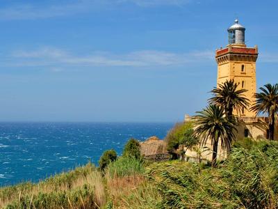 Leuchtturm an Küste mit blauem Meer, grünem Gras und Palmen unter klarem Himmel.