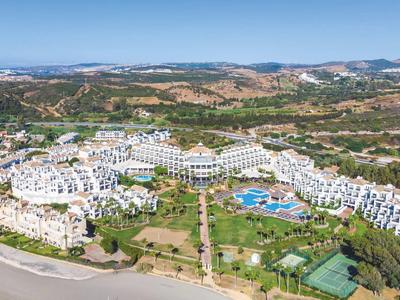 Grand complexe hôtelier blanc avec piscine et court de tennis dans un paysage vallonné.