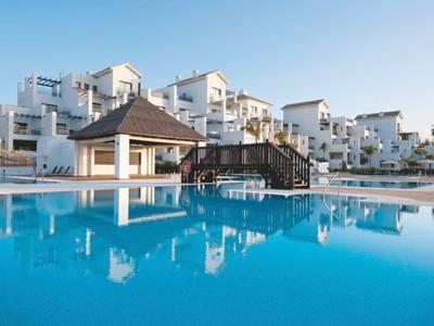 Großer blauer Pool mit Holzbrücke vor weißen Ferienhäusern unter blauem Himmel.