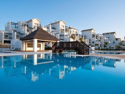 Hôtel avec grande piscine, pont en bois et bâtiments blancs à plusieurs étages sous un ciel clair.