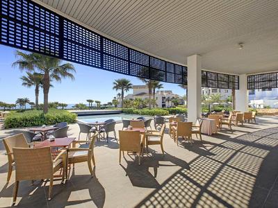 Terrasse couverte avec chaises et tables surplombant une piscine et des palmiers dans un hôtel.