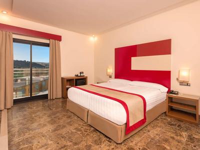 Hell eingerichtetes Hotelzimmer mit Doppelbett, rotem Kopfteilstoff und Balkon mit Aussicht.