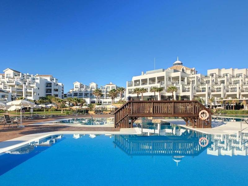 Großer blauer Pool vor weißen Hotels unter klarem Himmel mit Holzbrücke und Palmen.