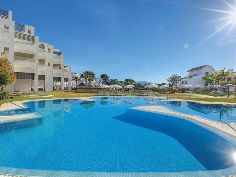 Großer blauer Pool mit weißem Treppenaufgang, umgeben von weißen Gebäuden und grünem Rasen unter blauem Himmel.