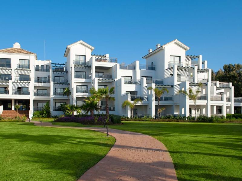 Weißes, mehrstöckiges Wohnhaus mit Balkon und grünem Rasen im Vordergrund bei blauem Himmel.