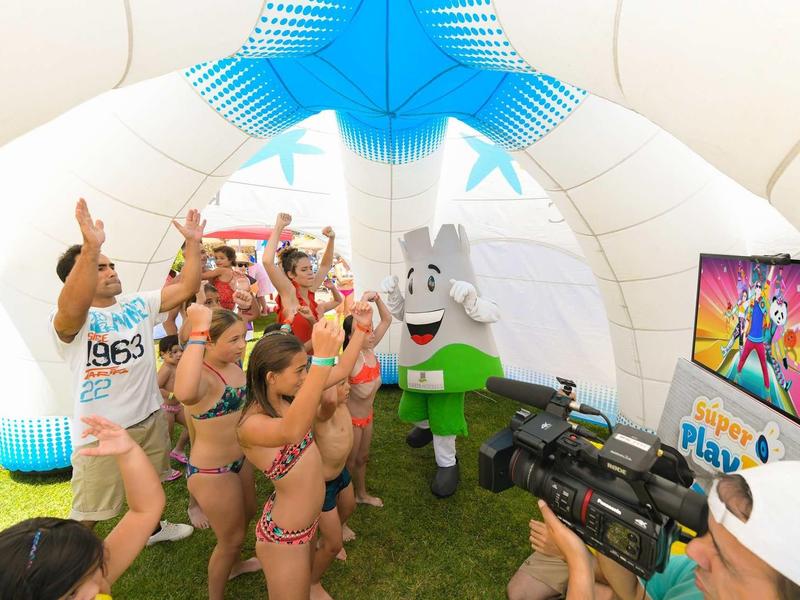 Kinder in Badebekleidung winken fröhlich vor einem aufblasbaren blau-weißen Spielplatz.