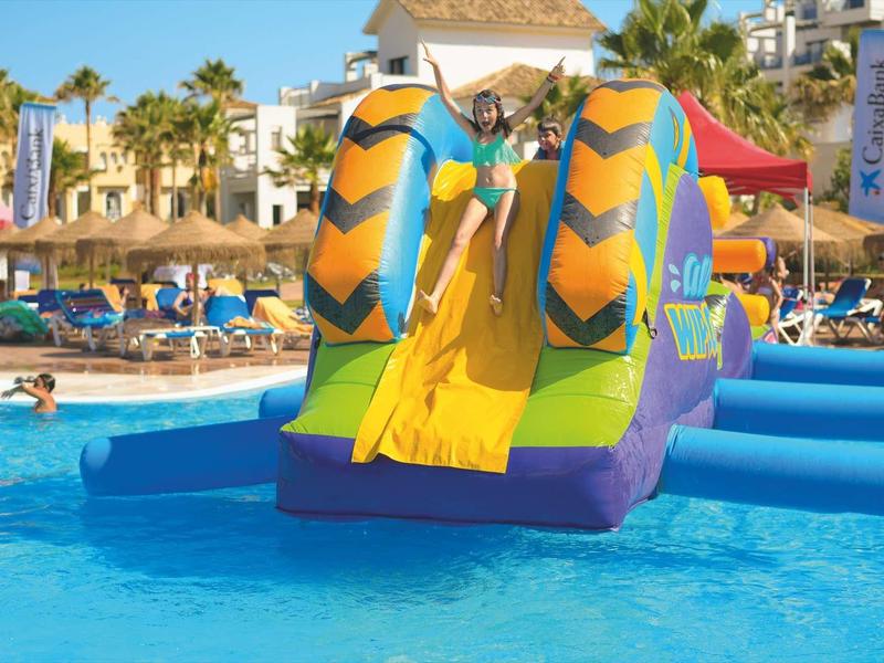 Kinder spielen auf bunter aufblasbarer Wasserrutsche im blauen Pool mit Palmen und Liegestühlen.