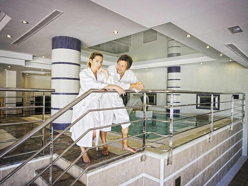 Zwei Personen in Bademänteln stehen neben einem Innen-Pool mit Edelstahlgeländer in modernem Raum.