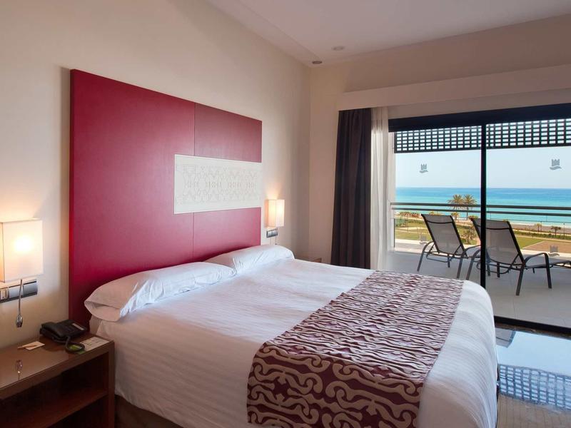 Modernes Hotelzimmer mit Doppelbett, rotem Kopfteil, Nachttischlampen und Meerblick vom Balkon.