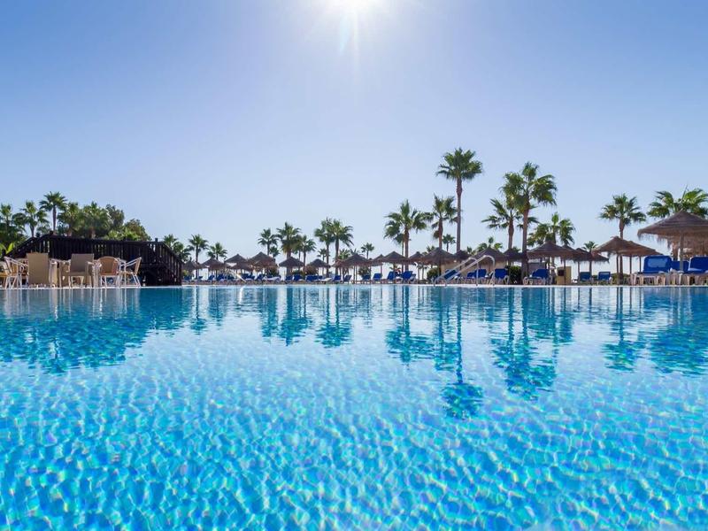 Großer blauer Pool mit Palmen, Sonnenschirmen und klarem Himmel im Hintergrund.