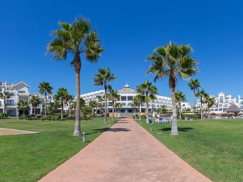 Große Hotelanlage mit weißen Gebäuden, Palmen und blauem Himmel, dahinter weitläufige Rasenflächen.
