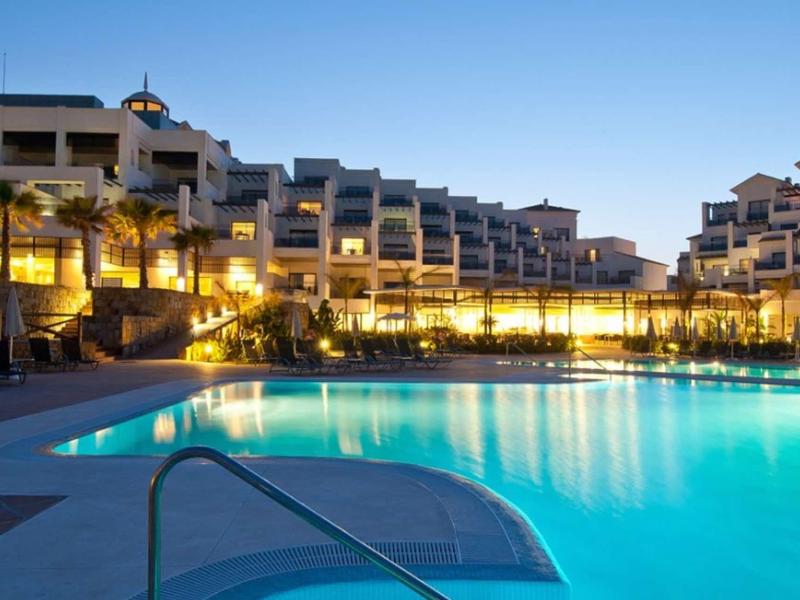 Großer, beleuchteter Pool vor modernen, mehrstöckigen Hotels bei klarem Abendhimmel.