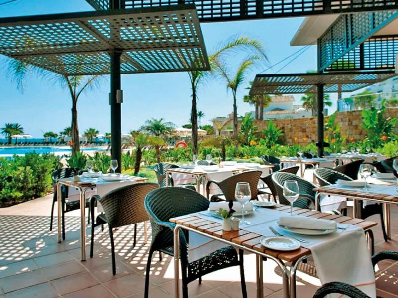 Freiluftrestaurant mit gedeckten Tischen, Stühlen, Palmen und Meerblick bei strahlendem Sonnenschein.