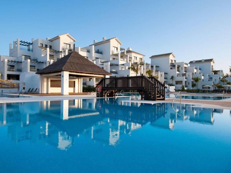 Großer blauer Pool vor weißen Gebäuden mit kleinen Terrassen und einem hölzernen Pavillon.