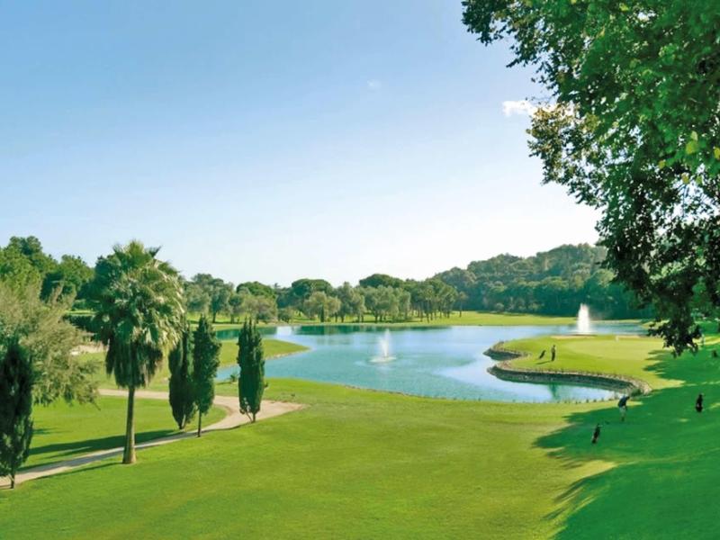 Grüner Golfplatz mit See, Bäumen und blauem Himmel im Hintergrund, klare und sonnige Atmosphäre.