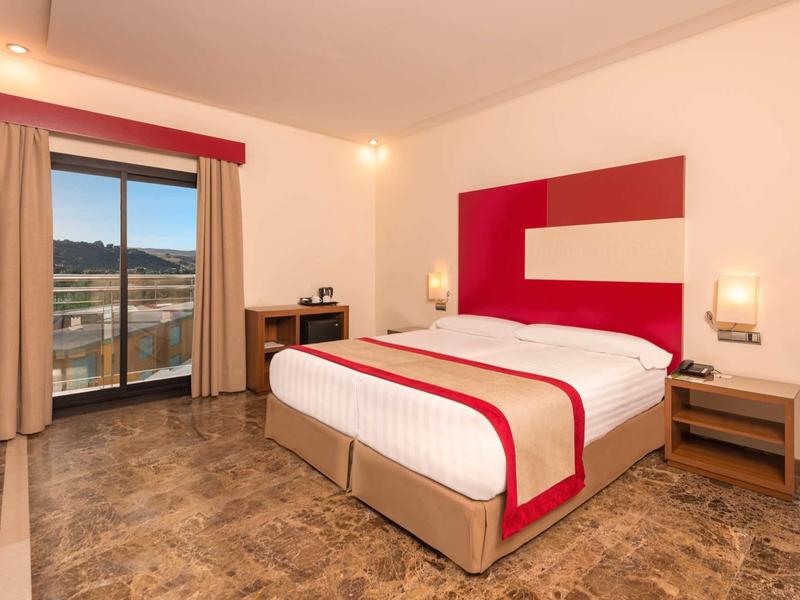 Hell eingerichtetes Hotelzimmer mit Doppelbett, rotem Kopfteilstoff und Balkon mit Aussicht.