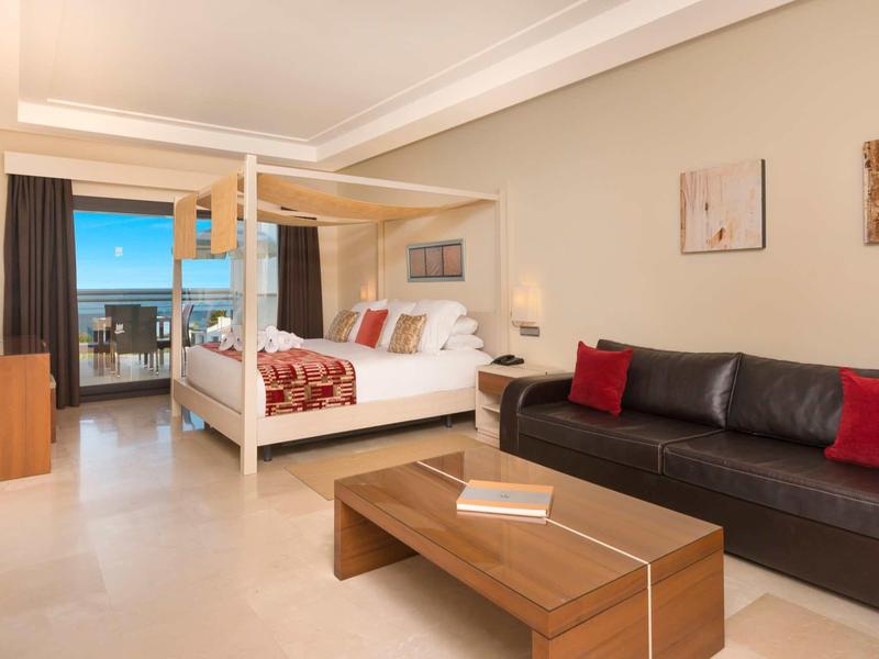 Geräumiges Zimmer mit Himmelbett, schwarzem Sofa, Holztisch und Blick auf Balkon mit Meer.