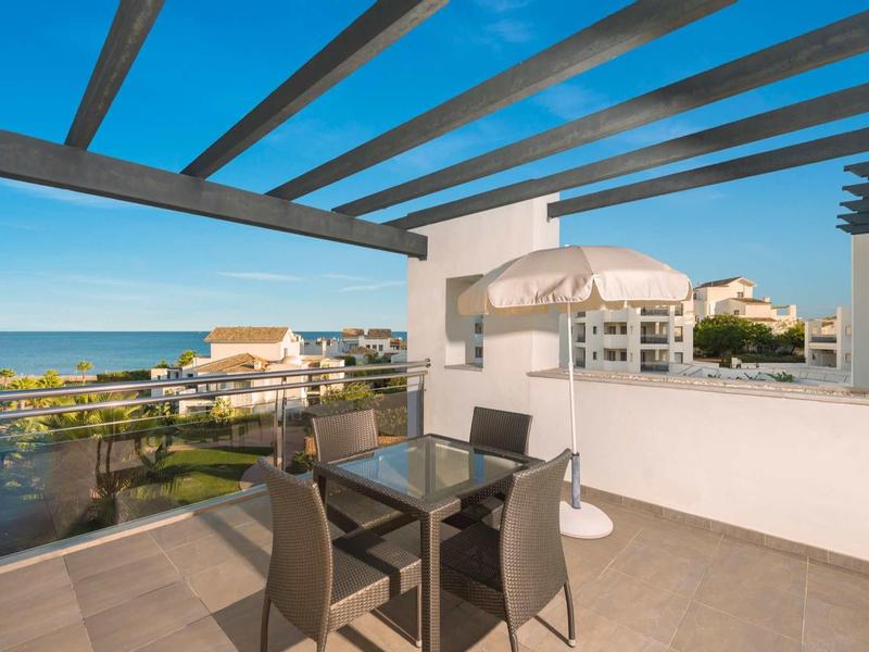 Terrassenbereich mit Glas-Gartentisch, vier Stühlen, weißer Wand und Meerblick bei blauem Himmel.