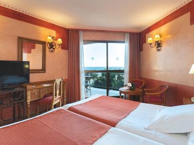 Helles Hotelzimmer mit Doppelbett, Fernseher, Schreibtisch, Sessel und Balkon mit Aussicht.