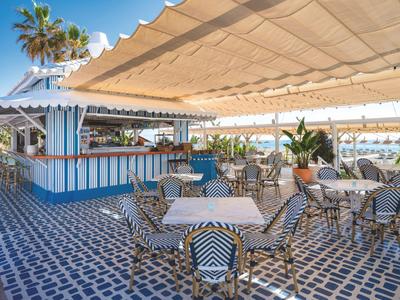 Offener Sitzbereich mit blauen und weißen Fliesen, Tischen und Stühlen unter einem großen Sonnendach.