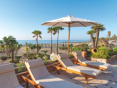 Sonnige Terrasse mit Liegestühlen, Sonnenschirm und Blick auf Palmen und Meer.