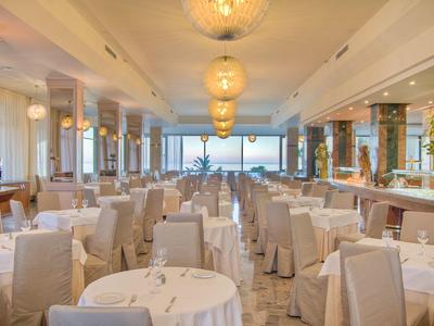 Großer heller Speisesaal mit weißen Tischdecken, Stühlen und runden Tischen, Blick auf Meer im Hintergrund.
