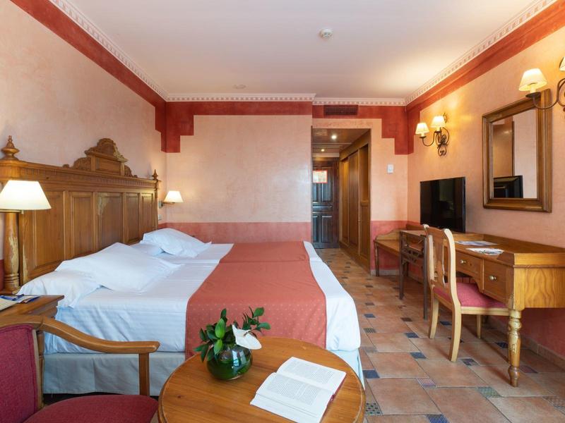 Ein helles Hotelzimmer mit großem Doppelbett, Schreibtisch, Stuhl und Pflanzen auf dem Nachttisch.