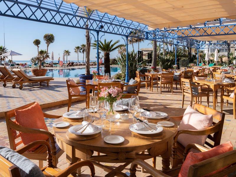Freiluftrestaurant am Pool mit Holzmöbeln, gedeckten Tischen und Blick auf Palmen und Wasserfläche.