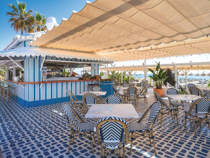 Offener Sitzbereich mit blauen und weißen Fliesen, Tischen und Stühlen unter einem großen Sonnendach.