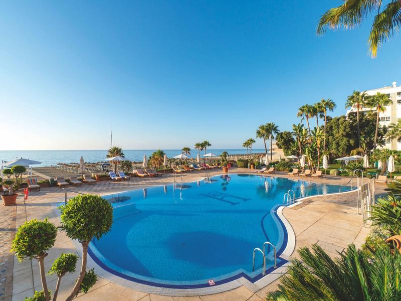 Großer, blauer Pool mit Palmen und Sonnenschirmen neben einem Hotel am sonnigen Strand.