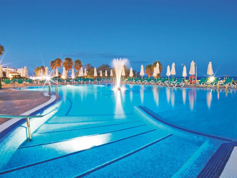 Großer blauer Pool mit Springbrunnen, weißen Liegestühlen und Palmen im Hintergrund bei Sonnenschein.
