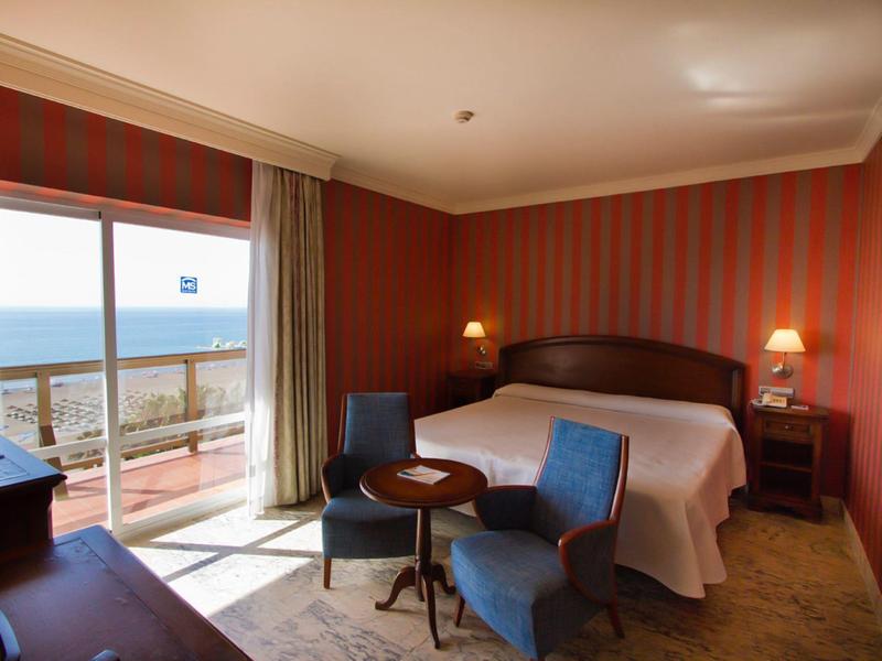 Hotelzimmer mit Doppelbett, blauen Sesseln, Nachtlampen und Balkon mit Meerblick bei Tageslicht.