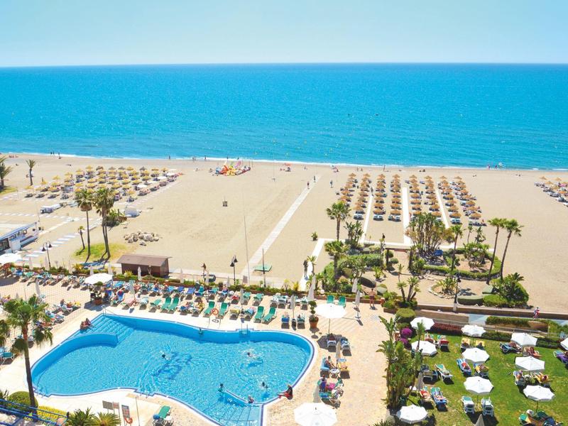 Blick auf einen Strand mit Sonnenschirmen, Liegen, Palmen und einem großen Pool im Vordergrund.