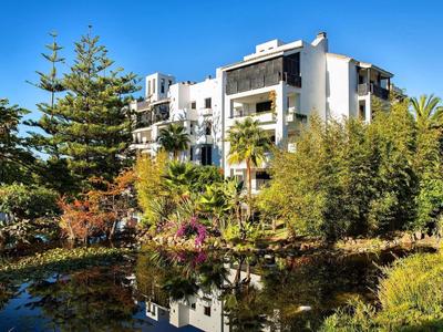 Modernes weißes Gebäude spiegelt sich im Teich, umgeben von Bäumen und grünem Buschwerk unter blauem Himmel.