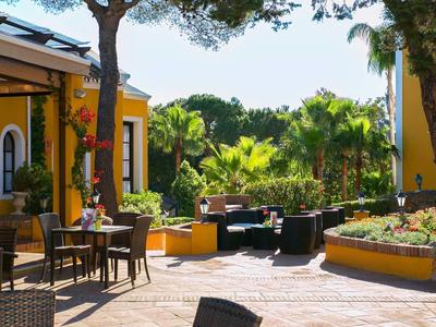 Ein sonniger Innenhof mit Bäumen, Tischen, Stühlen und bunten Gebäuden im Hintergrund.