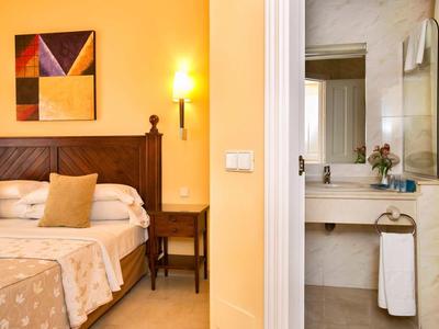 Schlafzimmer mit Holzbett, beige Wandleuchte, buntem Bild und angrenzendem Bad mit Waschbecken.