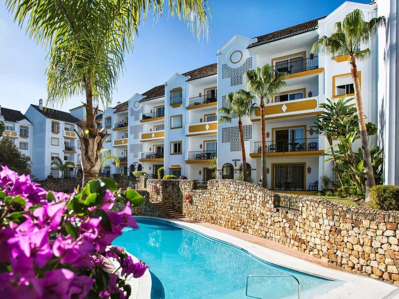 Wohnanlage mit Pool, Palmen und blühenden lila Blumen, weiß verputzte Gebäude und blauer Himmel.