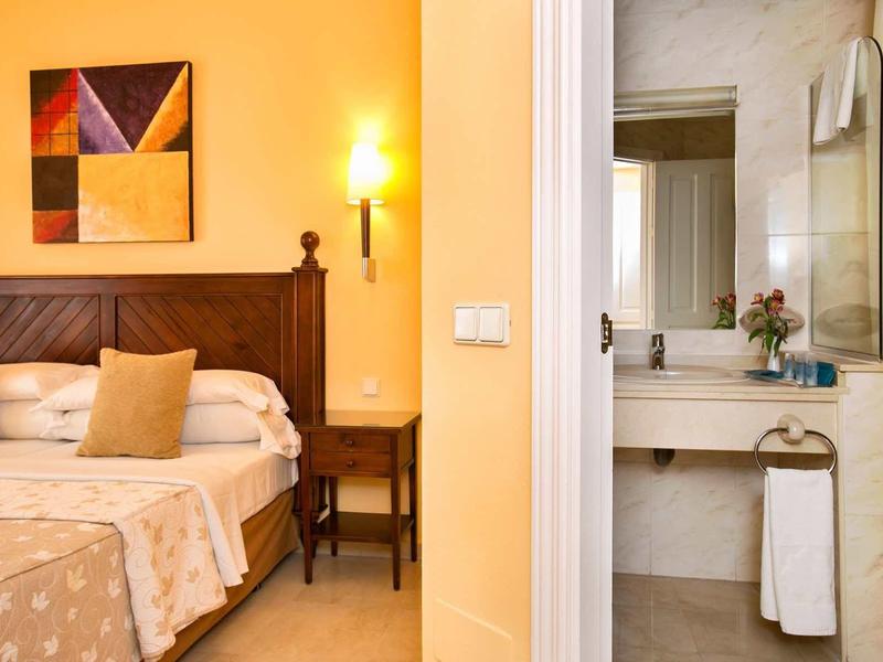 Schlafzimmer mit Holzbett, beige Wandleuchte, buntem Bild und angrenzendem Bad mit Waschbecken.