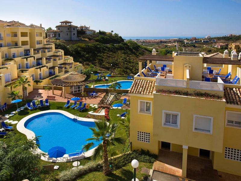 Mehrstöckige gelbe Wohngebäude umgeben einen Garten mit Pool und Liegestühlen, darüber blauer Himmel.
