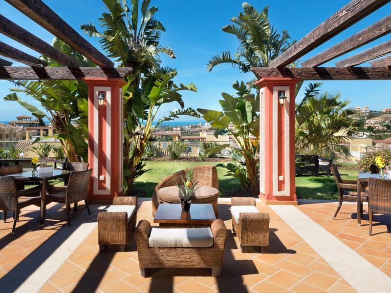 Terrasse mit Holzmöbeln, Pflanzen und Blick auf Garten und Meer bei klarem Himmel.